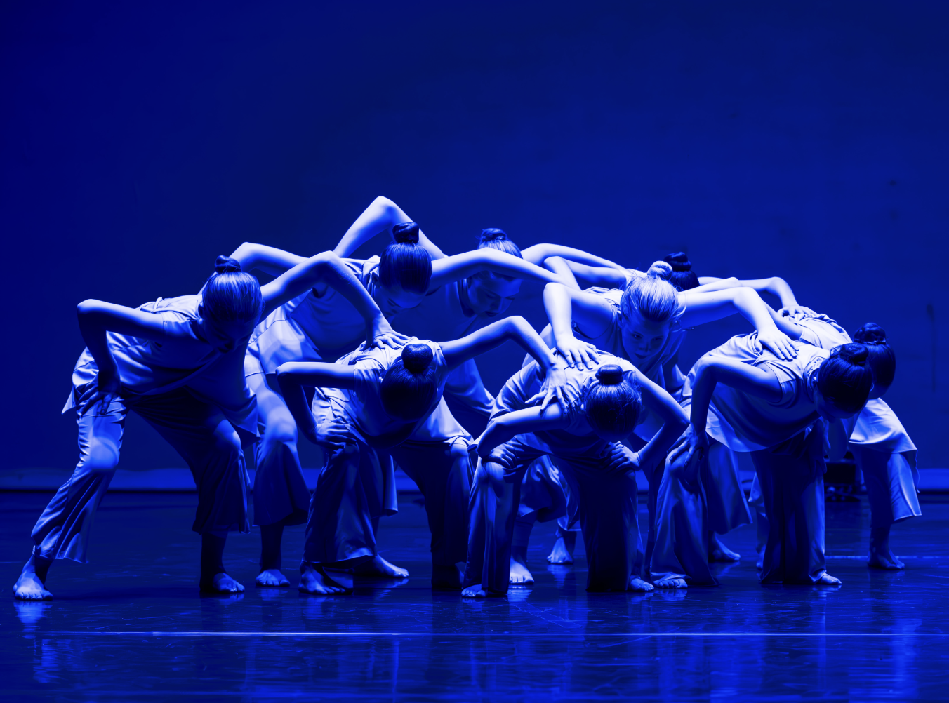 A group of dancers dance onstage with their arms around each other and their heads down in blue light.