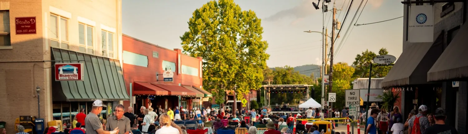 Image of a small downtown street festival with a stage and people in the street in front. Shops and stores line the street.