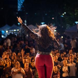 a woman in red pants and a denim jacket stands on a stage singing in front of a crown of people at night