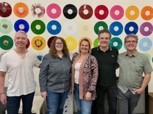 Five adults stand together indoors in front of a wall covered with colorful vinyl records arranged in a grid pattern, smiling at the camera, group photo style with casual clothing.