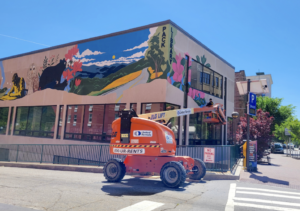 Orange construction lift in front of a building with a large, colorful mural of mountains, trees, and flowers on the facade.