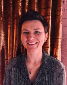a white woman with short dark hair smiles into the camera in front of a red curtain
