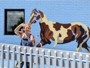 Smiling woman in denim overalls paints a brown-and-cream horse mural on a bright blue brick wall.