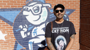 Man wearing sunglasses and a black cap poses in front of a colorful brick mural, wearing a t?shirt that reads 'World's Greatest Cat Mom' with a pictured figure on it.