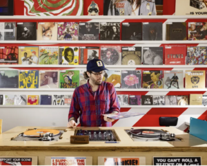 DJ in a red plaid shirt and cap stands at two turntables in a record store, examining a vinyl record with shelves full of colorful album covers behind him.