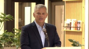 Man in a navy blazer and light blue shirt speaking at a podium with a microphone indoors, plants and wood paneling in the background.