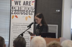 Woman at a lectern giving a presentation; banner behind reads 'WE ARE CONTINUOUS Learners' beside her.