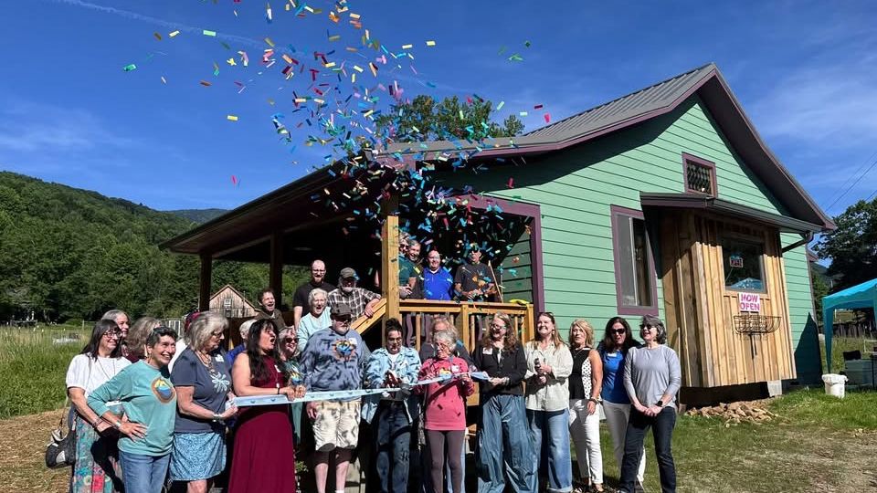 Group of people at a ribbon-cutting outside a small green wooden building as colorful confetti rains down on them