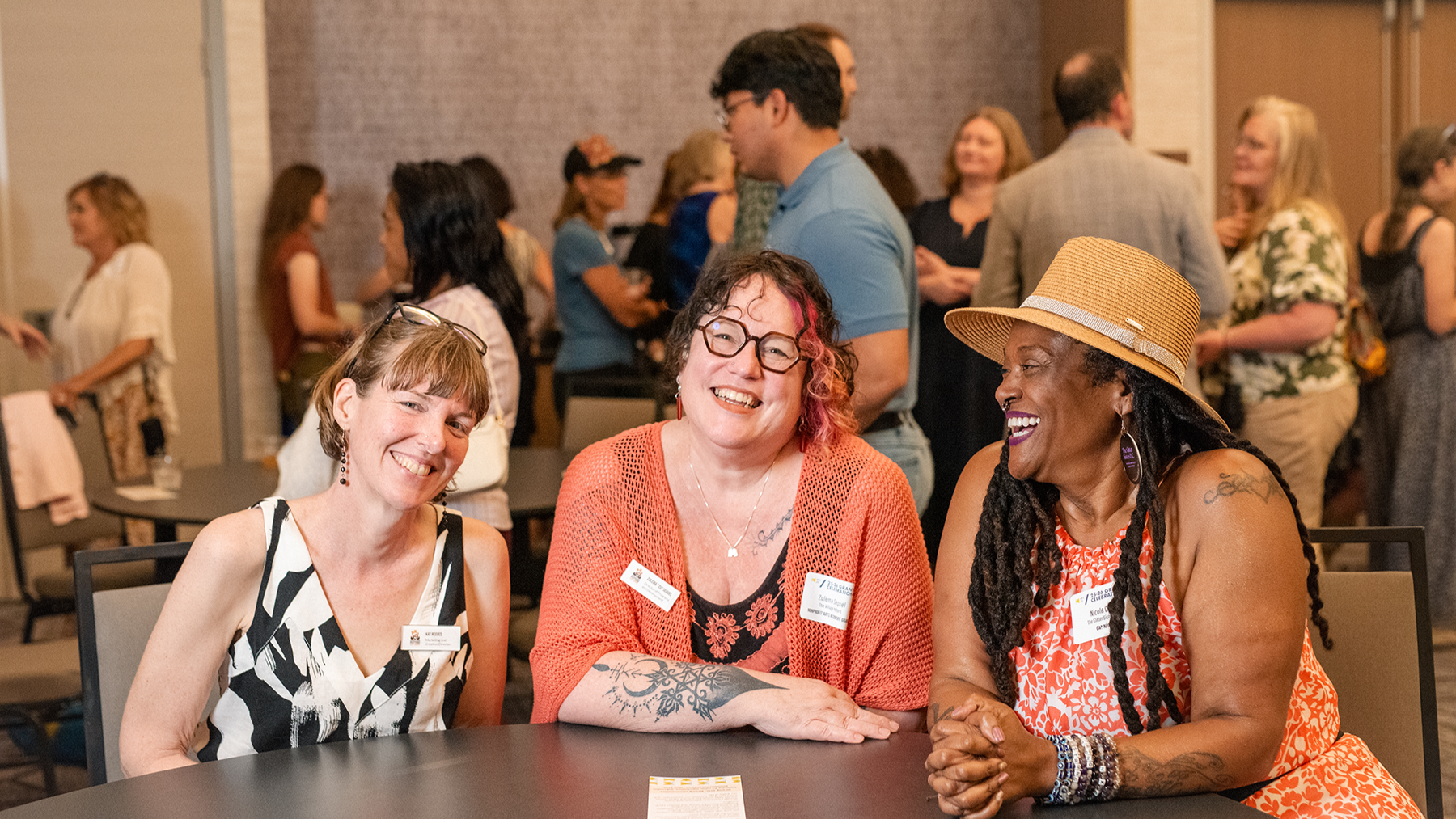 Three women sit at a table at a social event, laughing and chatting; each wears a name badge and casual attire.