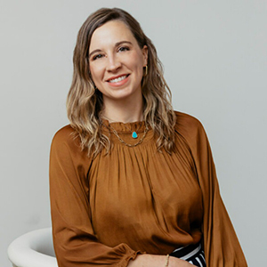 Smiling woman with wavy light brown hair wearing a brown satin blouse, posing against a light gray background on a white chair.