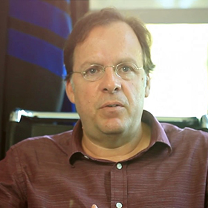 Middle-aged man with glasses wearing a maroon shirt, speaking indoors in an office setting.