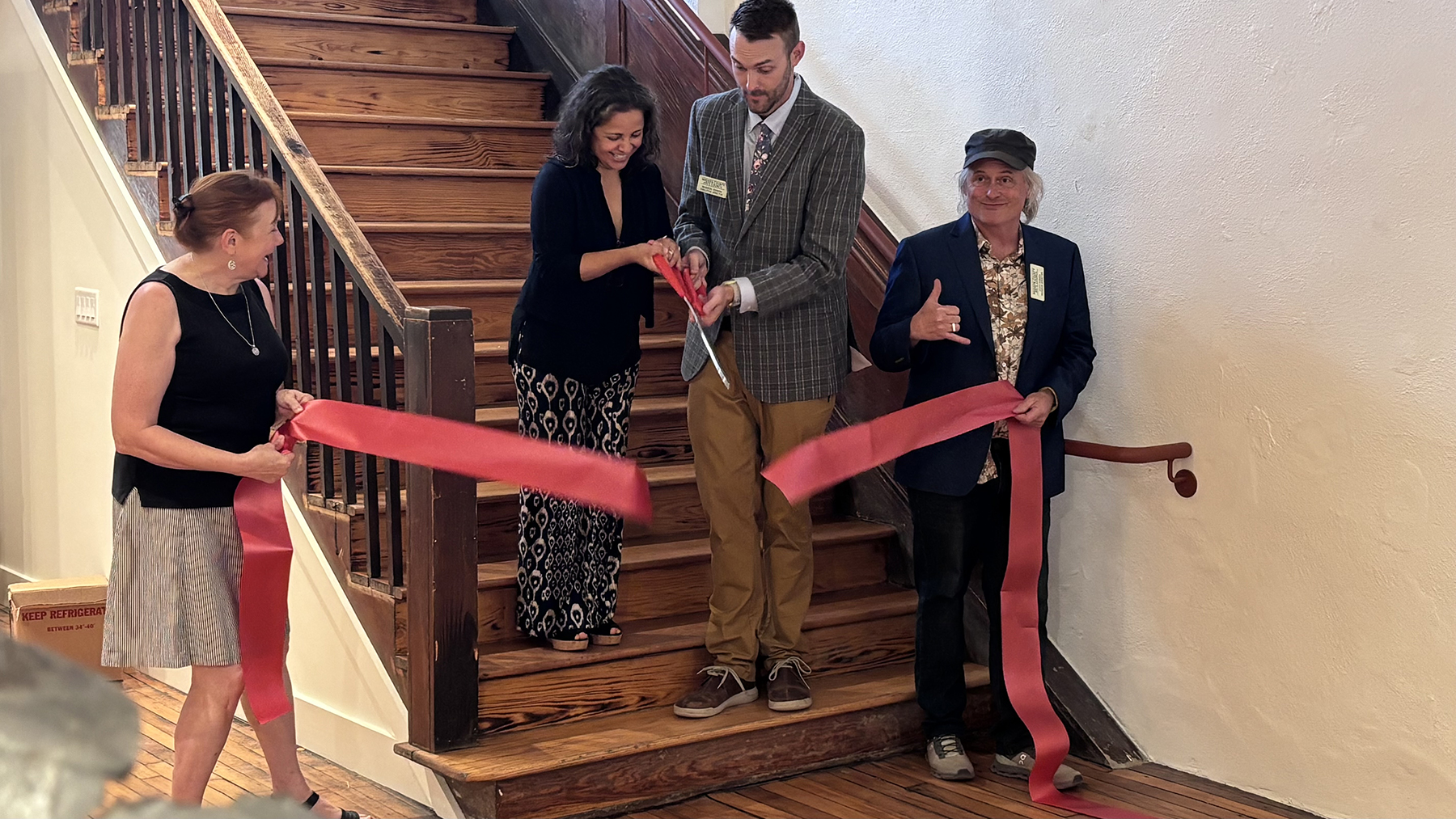 Four people participate in a ribbon-cutting ceremony on wooden stairs, as a man cuts the red ribbon with a pair of scissors nearby the others who watch and smile.