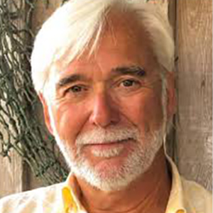 Smiling older man with white hair and beard, wearing a yellow shirt, outdoors beside a wooden fence.