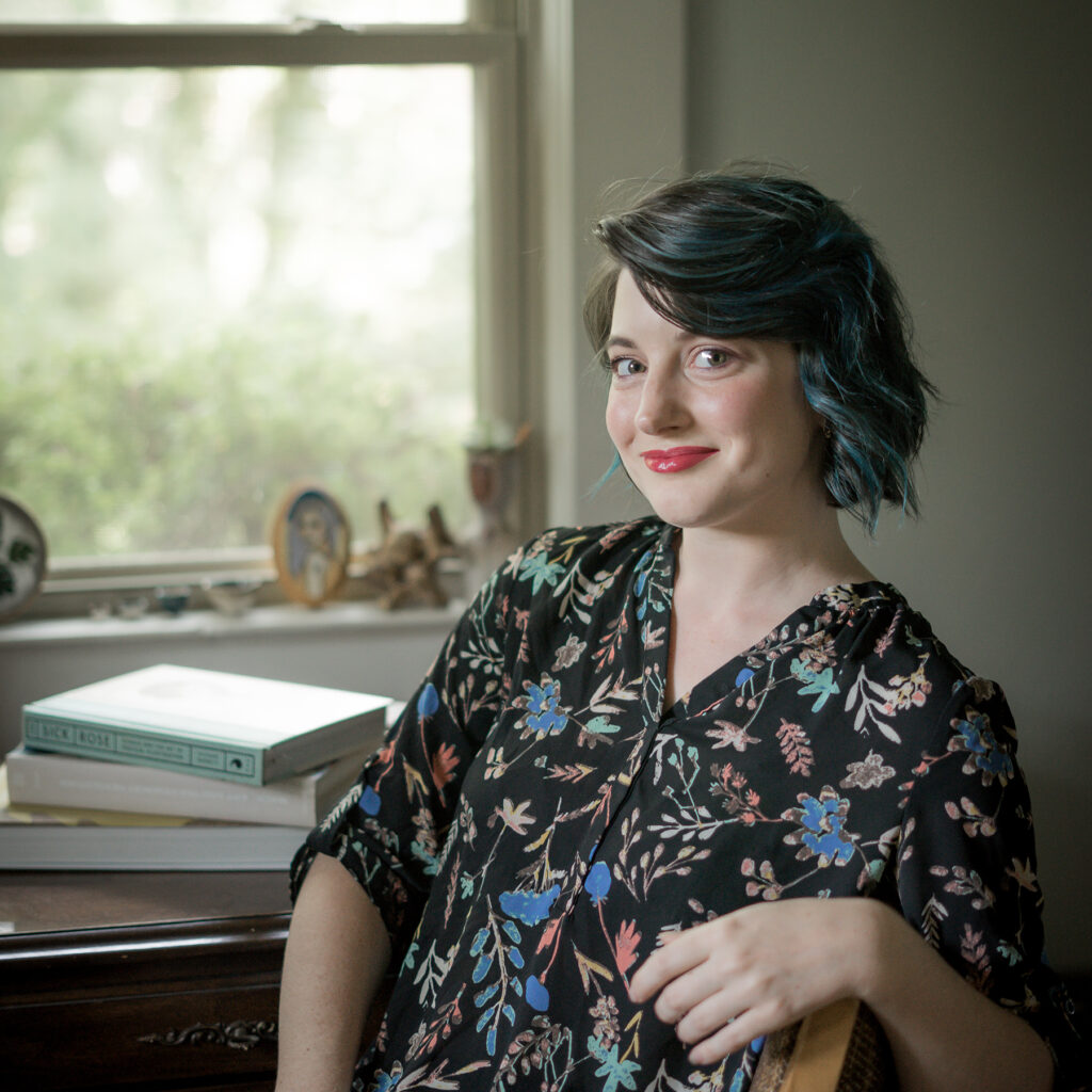 Smiling woman with teal-blue hair in a floral dress sits by a window with a piano and books in the background.
