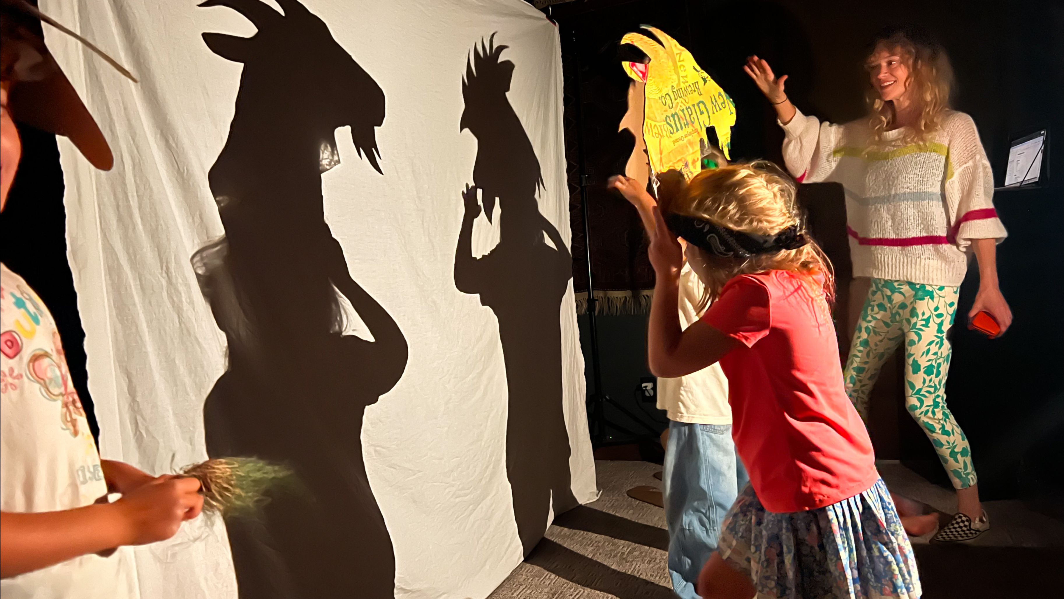 Children perform shadow puppetry behind a white screen, creating large black animal silhouettes; a smiling woman watches from the right.